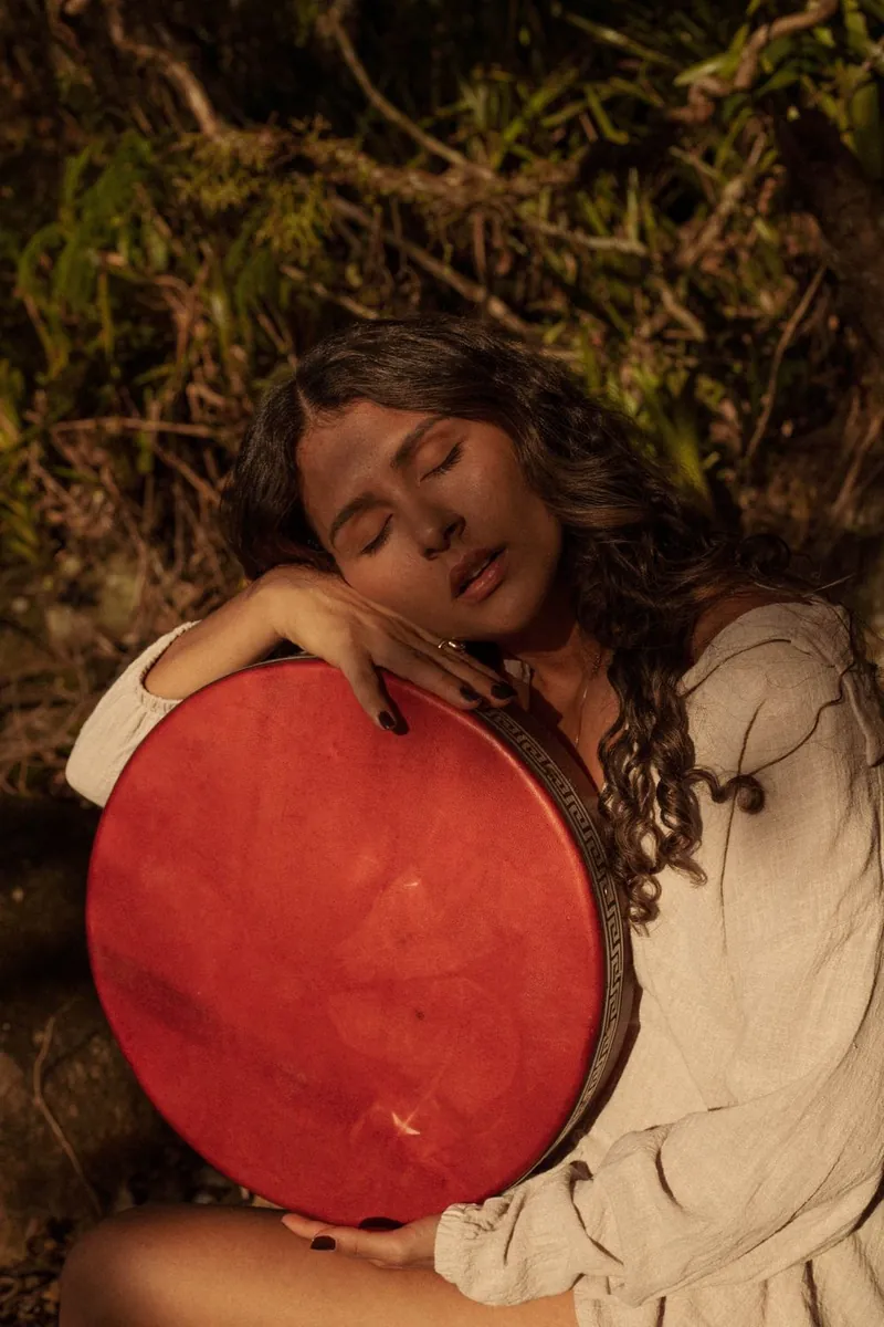Mulher de olhos fechados abraçando um tambor vermelho, em meio à natureza, na luz do fim da tarde